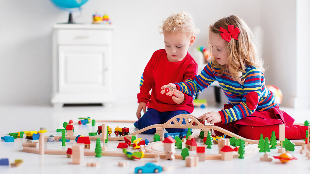 Zwei Kinder spielen zusammen mit einer Holzeisenbahn. Schienen, Häuser und Bäume sind auf dem Boden aufgebaut. Das eine Kind hält einen Waggon in der Hand, das andere greift danach.