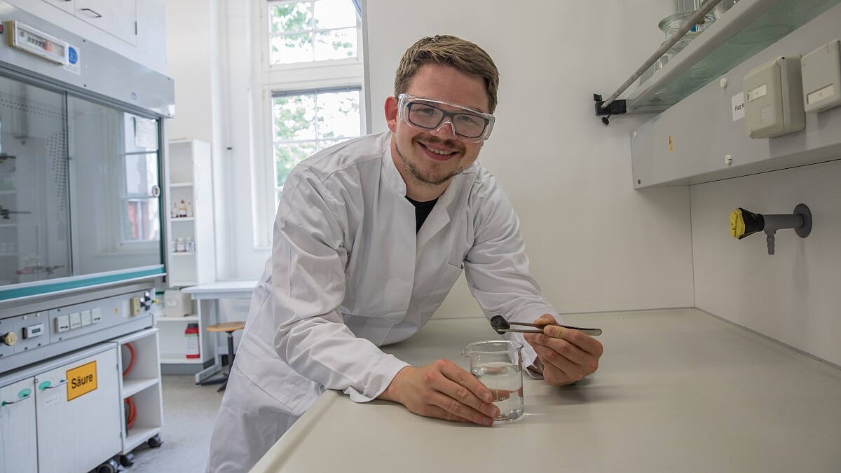 Ein Forscher steht lächelnd mit Schutzbrille und Kittel im Labor. Mit einer Pinzette hält er eine Materialprobe über einen Glasbecher mit Wasser.